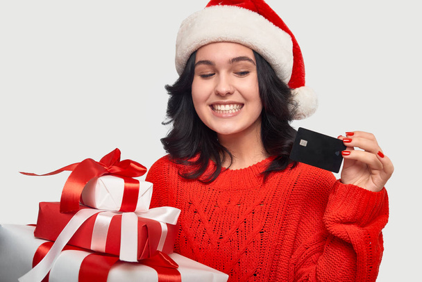 Mädchen in rotem Pullover und Weihnachtsmütze hält Geschenkboxen für Weihnachten in der Hand und zeigt ihre Bankkarte - Foto, Bild