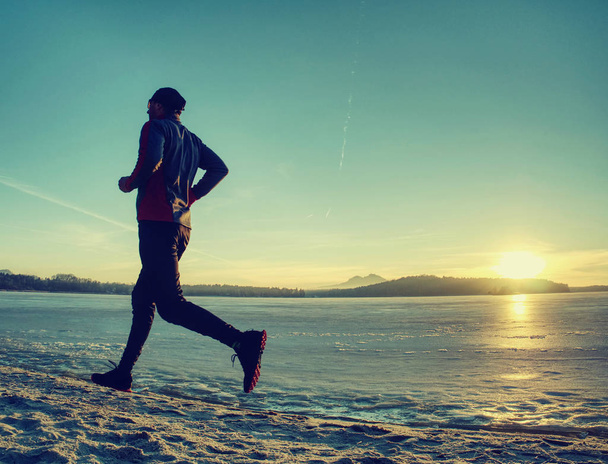 Urheilija juoksee rannalla jäätyneellä järvellä auringonlaskun aikaan.
 - Valokuva, kuva