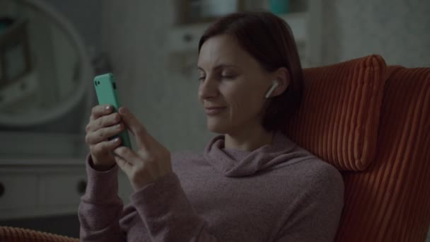Young female brunette adult with wireless earphones surfing online in smartphone sitting in cosy orange armchair in bedroom. Close up in slow motion.  - Filmmaterial, Video