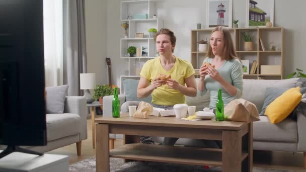 Jong stel eet een hamburger en kijkt TV in de woonkamer - Video