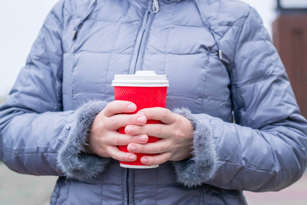 Młoda kobieta trzyma papierowy kubek z gorącym napojem. Czerwony kubek z białą plastikową pokrywą. Rozgrzej się na zewnątrz w mroźną pogodę. Nowy Rok i Święta Bożego Narodzenia. Podróż zimowa. - Zdjęcie, obraz