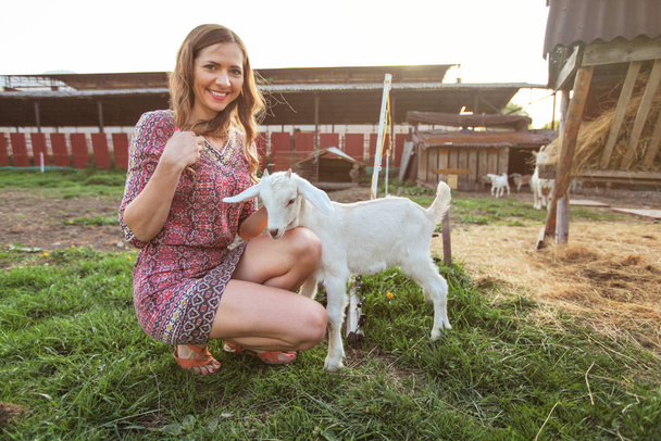 Young woman crouching, playing with goat kid, smiling, farm behi - Fotografie, Obrázek