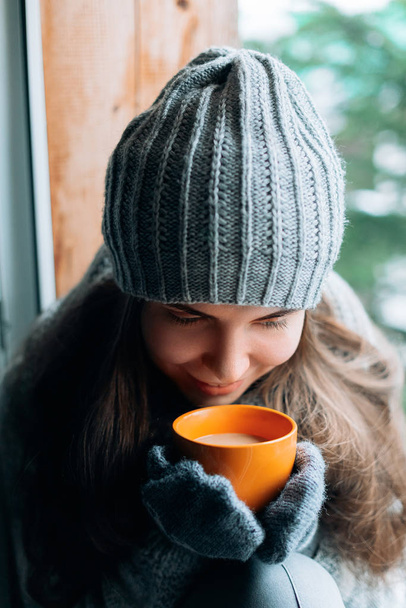 Mooie vrouw die een kop koffie of cacao vasthoudt en drinkt in handschoenen die thuis bij het raam zitten. wazig winter sneeuw boom achtergrond. Goedemorgen, gezelligheid en mensen concept  - Foto, afbeelding