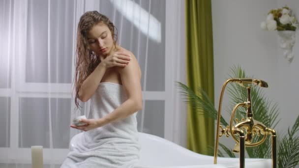 美容とボディケア女性の肘の痛みと健康管理の概念。窓の近くの白い居心地の良いアパートでベッドでリラックス化粧水クリーム保湿剤を適用する白人女性のスローモーション.  - 映像、動画