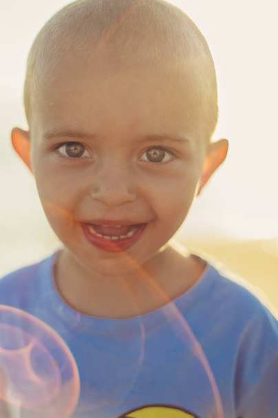 2-letá krátkosrstá holčička v modrém tričku s emocionálním výrazem na tváři při západu slunce. Světelné efekty ze slunce v kádru. Chlapec s velkýma očima a širokým úsměvem - Fotografie, Obrázek