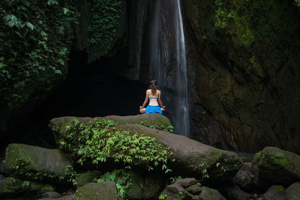 Young woman sitting on the rock, practicing yoga near waterfall. Hands in gyan mudra. Leke Leke waterfall, Bali. View from back. - Фото, зображення