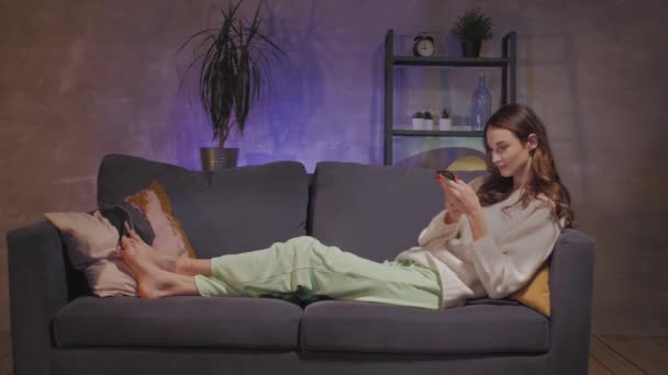 eine junge Frau sitzt auf einem Sofa in einem gemütlichen Zimmer schreibt Nachrichten am Telefon und ist enttäuscht - Filmmaterial, Video