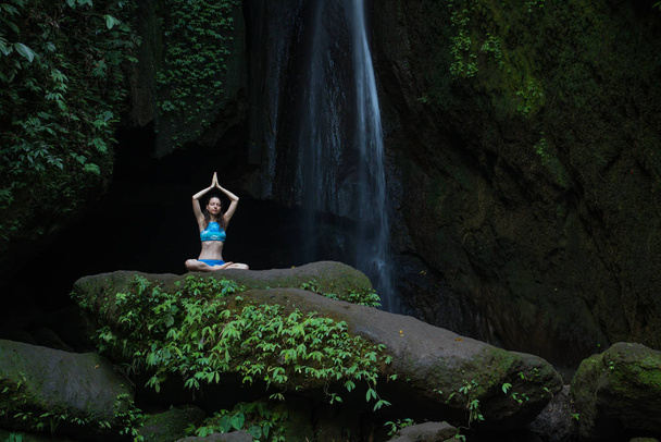 Young Caucasian woman meditating, practicing yoga at waterfall. Hands raising up in namaste mudra. Leke Leke waterfall, Bali, Indonesia.  - Photo, Image