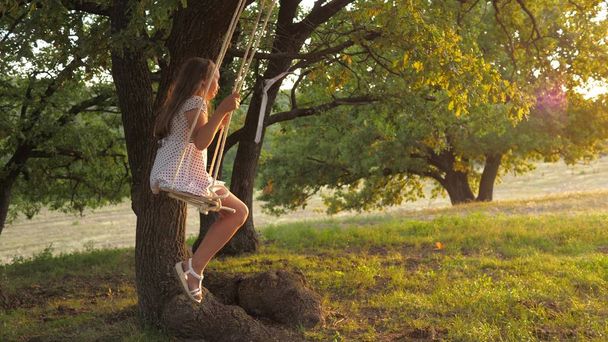 child swinging on a swing in park in sun. young girl swinging on rope swing on an oak branch. teen girl enjoys flight on swing on summer evening in forest. concept of happy family and childhood. - Foto, afbeelding