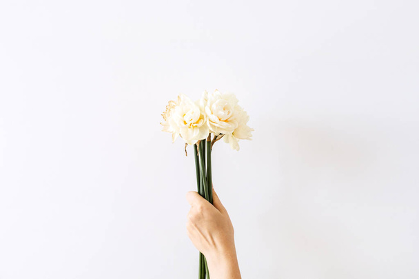 Female hand hold narcissus flowers bouquet on white background. Flat lay, top view minimal floral composition. - Fotó, kép