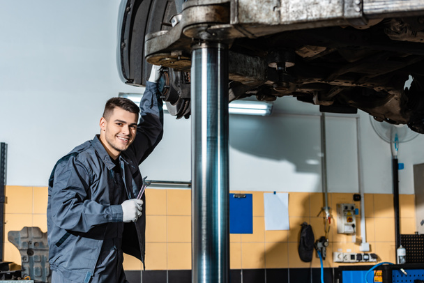 smiling mechanic holding wrench and looking at camera while standing under car raised on car lift - Foto, Bild