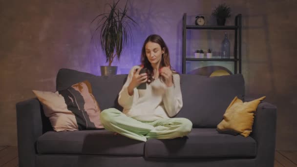 Nuori nainen istuu sohvalla viihtyisässä huoneessa ottaa selfien ja hymyilee
 - Materiaali, video