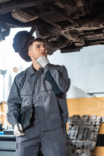thoughtful mechanic holding hand near face while looking at car bottom - 写真・画像