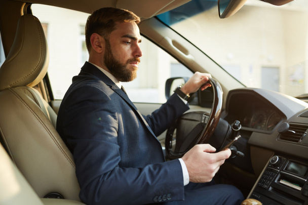 Young businessman sitting behind the steering wheel and driving while reading message on mobile phone - Foto, Imagen
