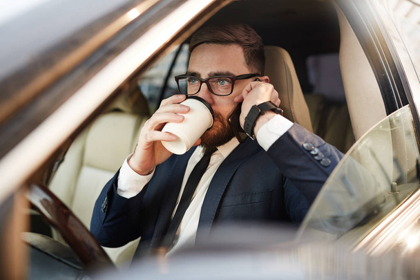 Young businessman in eyeglasses drinking coffee and talking on mobile phone in the car - Fotoğraf, Görsel