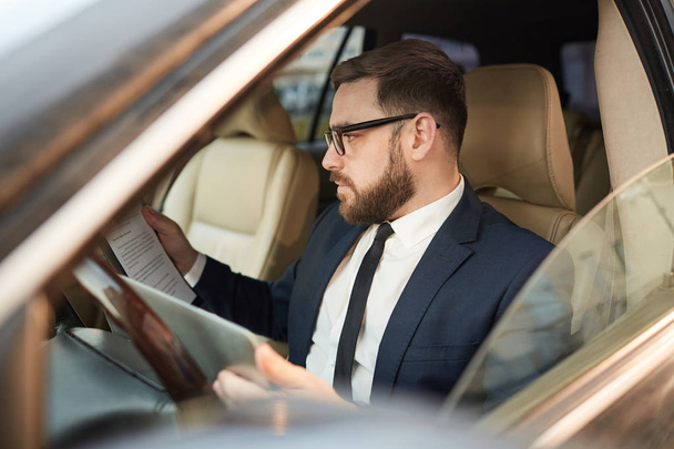 Jeune homme d'affaires barbu portant des lunettes, assis dans la voiture et examinant un contrat d'entreprise - Photo, image