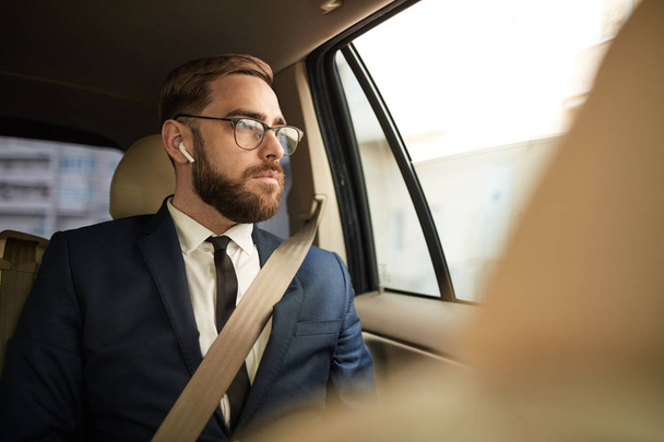 Νεαρός επιχειρηματίας με γυαλιά ηλίου που κάθεται στο πίσω κάθισμα και κοιτάζει μέσα από το παράθυρο με στοχαστικό θέαμα κατά τη διάρκεια της διαδρομής με ταξί - Φωτογραφία, εικόνα