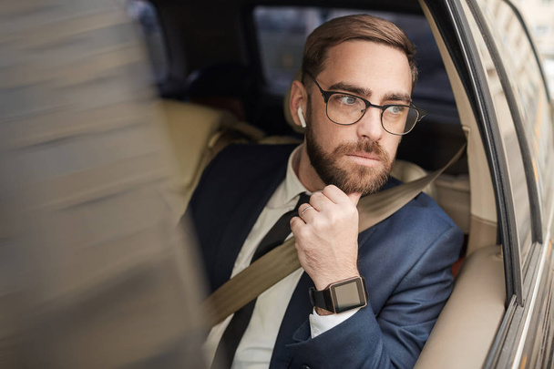 Νεαρός στοχαστικός επιχειρηματίας με γυαλιά που κάθεται στο πίσω κάθισμα του ταξί και κοιτάζει μέσα από το παράθυρο - Φωτογραφία, εικόνα