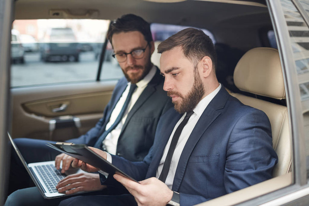 Two business partners using laptop and discussing documents together on the back seat during driving  - 写真・画像