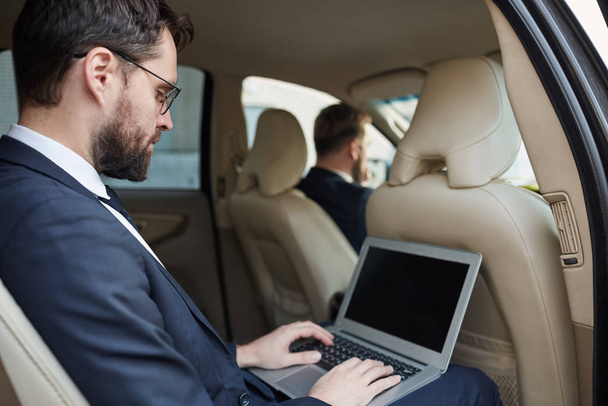Молодий бізнесмен у костюмі сидить на задньому сидінні автомобіля і працює онлайн за допомогою ноутбука під час водіння автомобіля
 - Фото, зображення