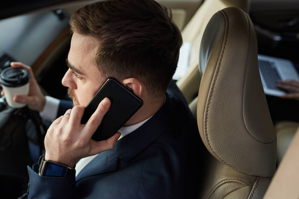 Молодой бородатый бизнесмен разговаривает по мобильному телефону и пьет кофе, сидя за рулем в машине - Фото, изображение
