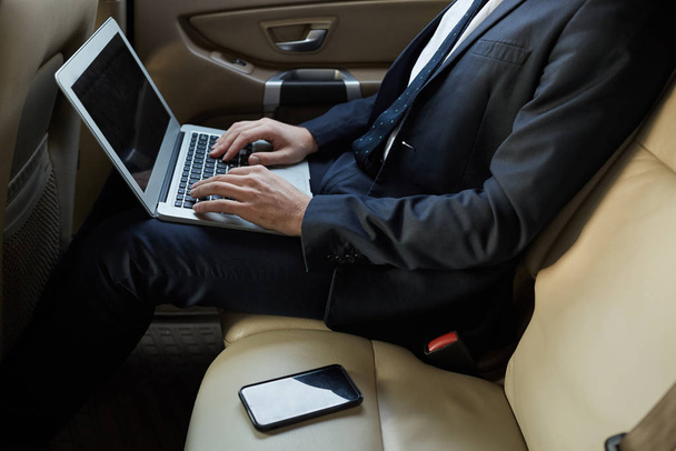 Gros plan d'un homme d'affaires en complet assis sur le siège arrière d'une voiture de luxe et tapant sur un ordinateur portable travaillant en ligne pendant la conduite - Photo, image