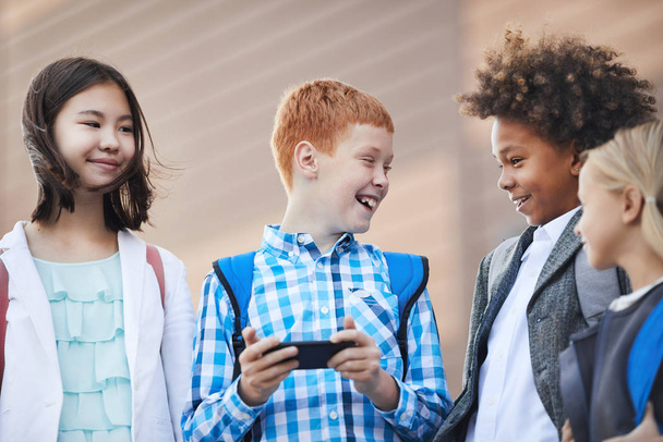 Niño cabellos rojos de pie con teléfono móvil y risas mientras hablan con compañeros de clase al aire libre. - Foto, Imagen