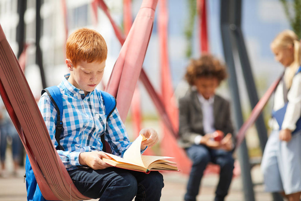 Школьник с рюкзаком сидит на качелях и читает книгу на школьной детской площадке - Фото, изображение