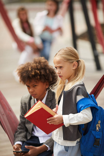 Blond schoolgirl with backpack behind back holding book standing on the playground with African boy using mobile phone - Foto, Bild