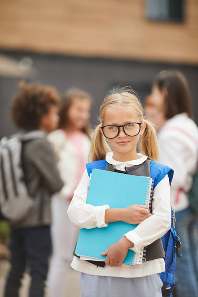 Porträt eines blonden Mädchens mit Brille, das Schulbücher hält und in die Kamera schaut, während es draußen mit Klassenkameraden im Hintergrund steht - Foto, Bild