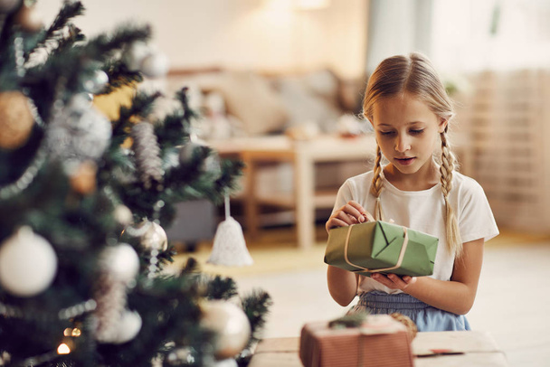 Little blonde girl holding gift box in hands and reading postcard while sitting on the floor near the Christmas tree - Photo, Image