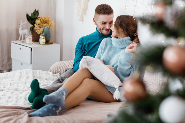 Boldog pár karácsonyi dekorációban otthon. Szilveszter este, díszített fenyő. Téli nyaralás és szerelem koncepció.Fiatal boldog pár ölelése és pihentető kényelmes kanapén. - Fotó, kép