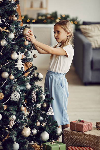 Klein meisje met blond haar staande en versieren kerstboom met speelgoed in de woonkamer thuis - Foto, afbeelding