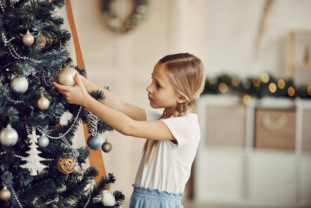 Niedliches kleines Mädchen steht zu Hause am Weihnachtsbaum und untersucht Spielzeug - Foto, Bild