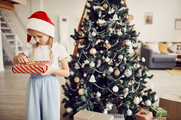 Маленька дівчинка в капелюсі Санта тримає подарункову коробку в обгортковому папері і читає листівку, стоячи біля ялинки вдома
 - Фото, зображення