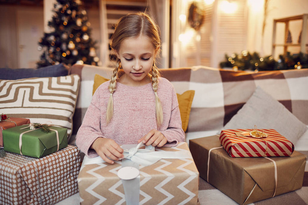 Niña sentada en el sofá y abriendo grandes regalos de Navidad que recibe de Santa Claus para Navidad en casa
  - Foto, Imagen