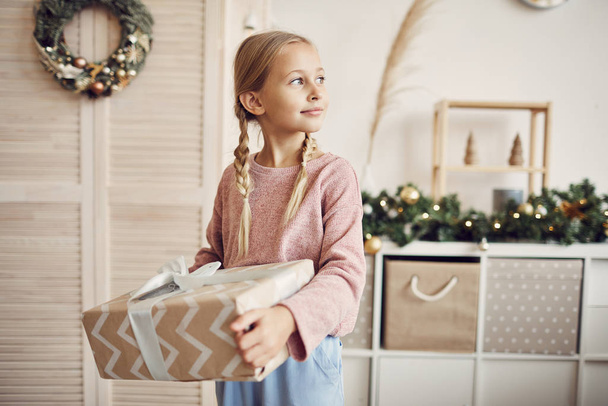 Petite fille aux cheveux blonds tenant un gros cadeau dans les mains et allant le long de la pièce à la maison
 - Photo, image