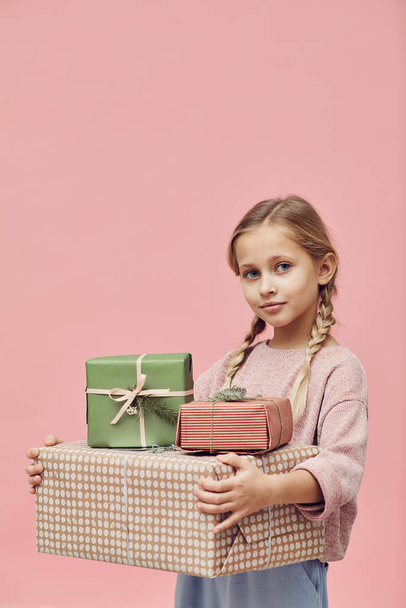 Portrait of little girl with blond hair holding wrapped gift boxes and looking at camera over pink background  - Zdjęcie, obraz