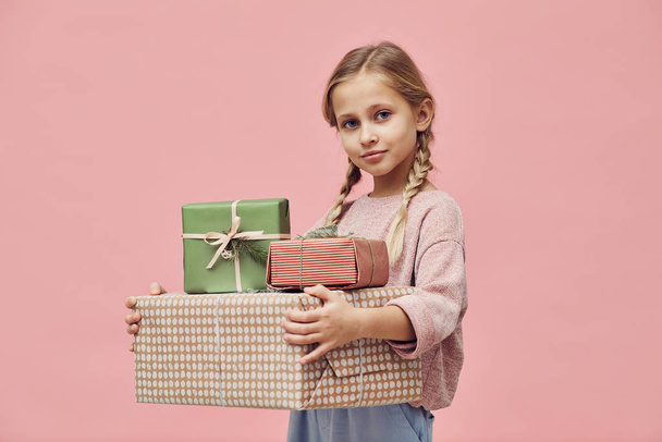 Portrait of little girl with blond hair holding wrapped gift boxes and looking at camera over pink background  - Foto, afbeelding