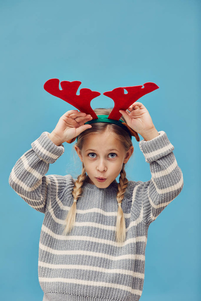 Portret van een grappig klein meisje dat hertengewei draagt en doet alsof er een kersthert tegen de blauwe achtergrond staat  - Foto, afbeelding