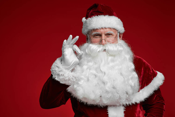 Porträt des Weihnachtsmannes mit weißem Bart, der ein Ok-Zeichen setzt und in die Kamera blickt - Foto, Bild