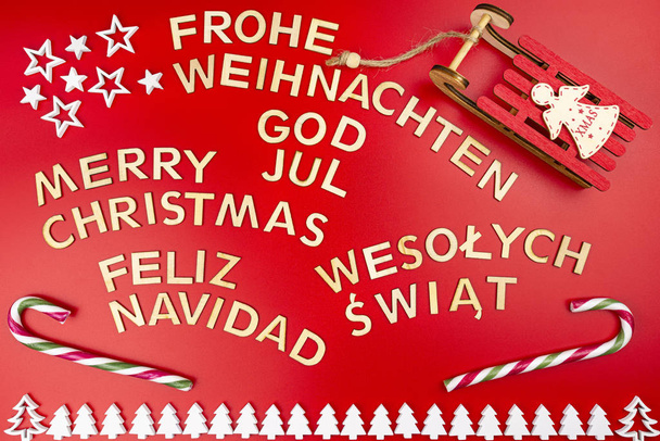 Inscription Merry Christmas in German, English, Polish, Swedish and Spanish, lying flat from above. Made of wooden inscriptions, isolated on a red background, with visible sledges and candy canes. - 写真・画像