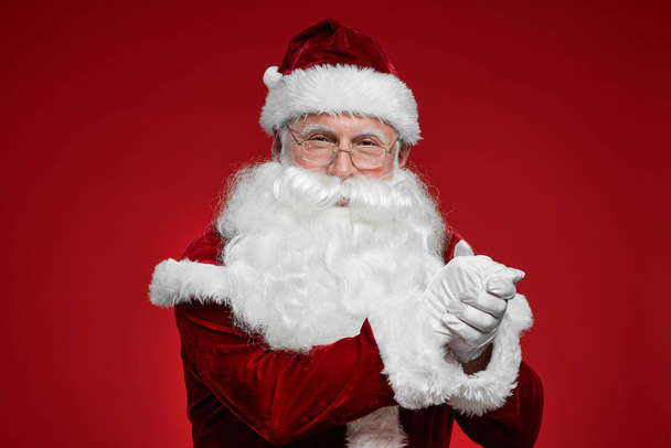 Portrait of Santa Claus in eyeglasses and with white beard showing respect and looking at camera on red background - Φωτογραφία, εικόνα