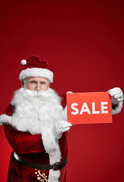 Portrait of Santa Claus in costume and with white beard holding sale sign against the red background - Fotó, kép