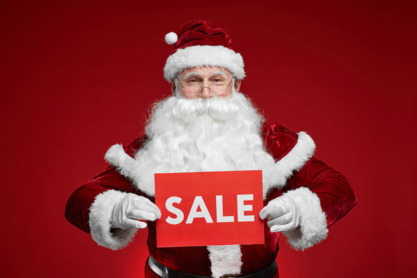 Portrait of Santa Claus holding sale sign and smiling at camera against the red background - Foto, Bild