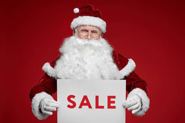 Portrait of Santa Claus holding placard with sale sign and looking at camera over red background - Foto, immagini