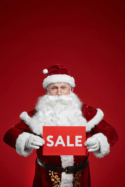 Portrait of senior man with white beard and in Santa costume showing sale sign on red background - Photo, Image