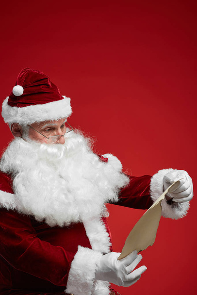 Santa Claus wearing eyeglasses standing and reading letter in hands against the red background - Photo, Image