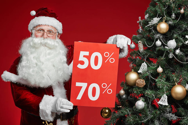 Portrait of senior man in Santa costume holding placard with big sale while standing near the Christmas tree over red background - Photo, Image