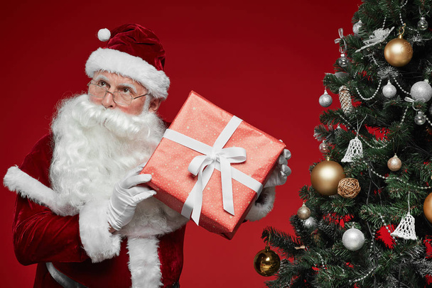 Père Noël tenant boîte cadeau enveloppé dans du papier d'emballage rouge tout en se tenant près de l'arbre de Noël sur fond rouge
 - Photo, image